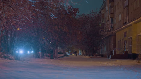 residential buildings and snow-covered street during winter evening with blurred figures walking in the distance, car headlights illuminating through frosty trees