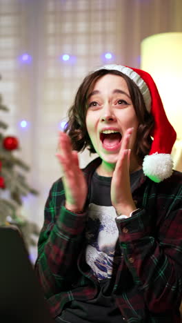 beautiful brunette lady in santa cap looks surprised on the laptop. girl rejoices clapping hands happily. close up. vertical video.