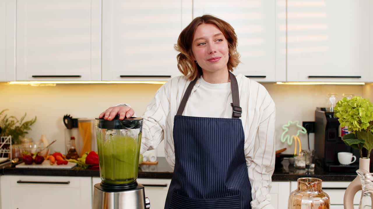 Giovane donna sorridente che prepara un frullato verde salutare nel frullatore per la dieta, in una accogliente cucina di casa.