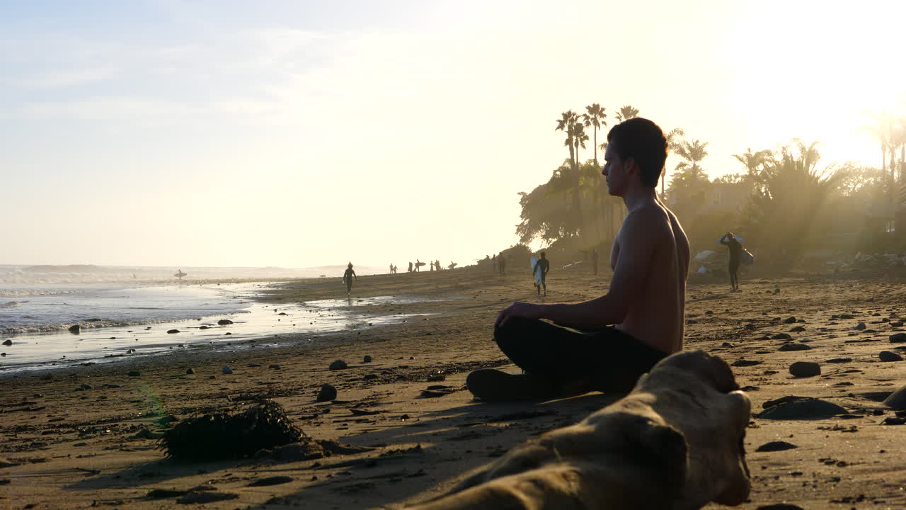 un hombre se sienta en una pose de meditación para aliviar el estrés en una playa de puesta de sol con palmeras y personas en silueta en la orilla del océano