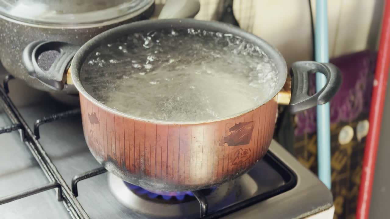 Close-up of water rapidly bubbling in a metal saucepan on a gas stove with blue flame and steam, indoor kitchen cooking scene showing heat and boiling process