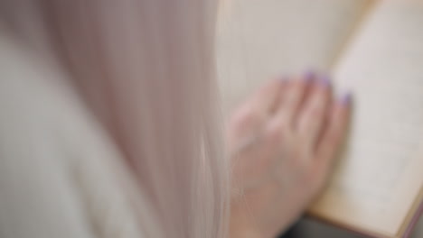 blurry side view of woman with long pastel hair seated on mall bench hands gently turning pages of hardcover novel soft focus background shows shoppers on moving walkway creating calm reading moment