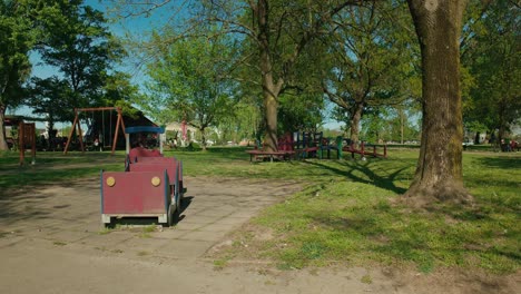 Red-children's-train-sits-idle-by-playground-swings-in-the-vibrant-greenery-of-Jarun-Lake-Park,-Zagreb-Croatia