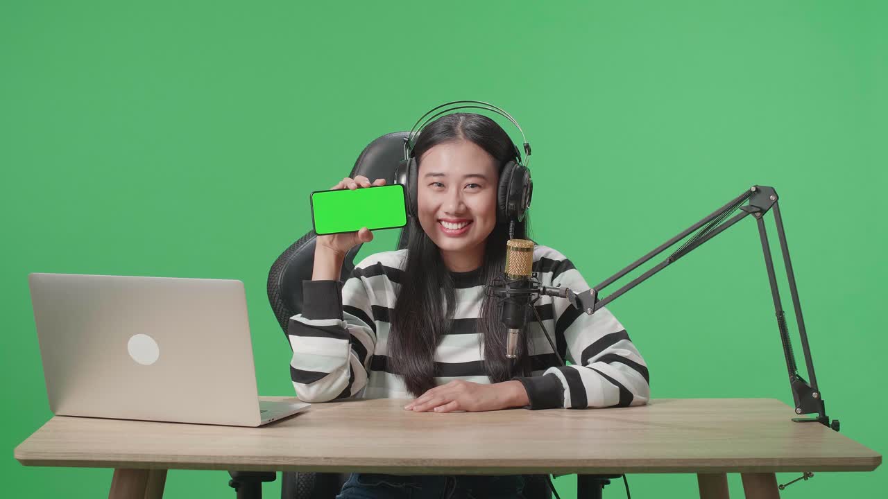 A Smiling Asian Woman With Headphone And Computer Showing Green Screen Mobile Phone While Sitting In Front Of Green Screen Background