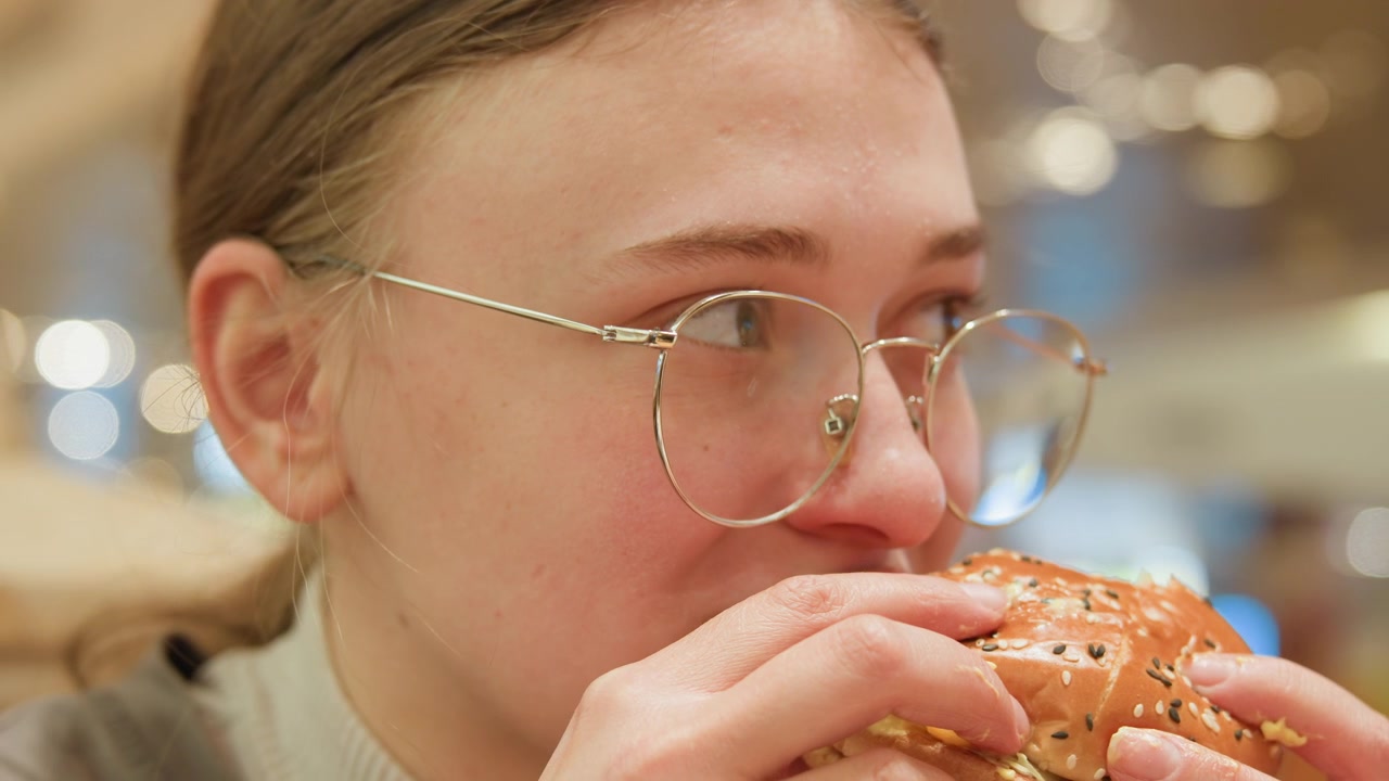 White traveler tasting local burger with glasses slow contemplative chew and subtle smile, seeded bun held close with crumbs on chin, warm food court lighting and cozy evening vibe