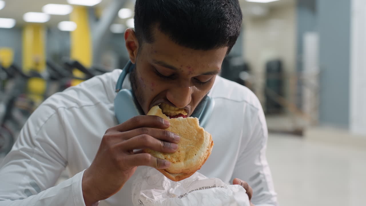 young man wearing white gym wear and headphones takes bite of sandwich while seated in modern fitness center with blurred exercise equipment in background after workout session
