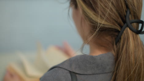 close up back view of a lady in grey clothing flipping through a book as the wind gently blows the pages, her hair is tied with a black clip, and the background is softly blurred