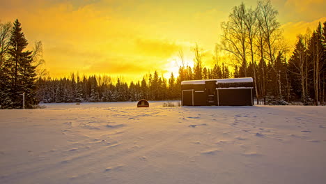 Hermosa-Puesta-De-Sol-Amarilla-Sobre-Un-Campo-De-Bosque-De-Coníferas-De-Invierno,-Con-Una-Cabaña-Y-Sauna-De-Barril