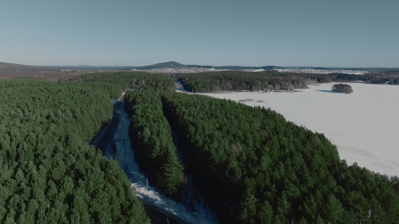Drönarfilm av en slingrande väg genom tät tallskog bredvid en vidsträckt frusen sjö en klar vinterdag i Uralbergen, Ryssland, ett naturskönt vildmarkslandskap.