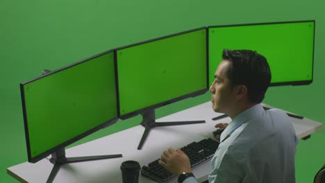 vista lateral de un hombre asiático pensando en algo y luego levantando su dedo índice mientras trabaja con un simulacro de varios monitores de computadora en un estudio de pantalla verde