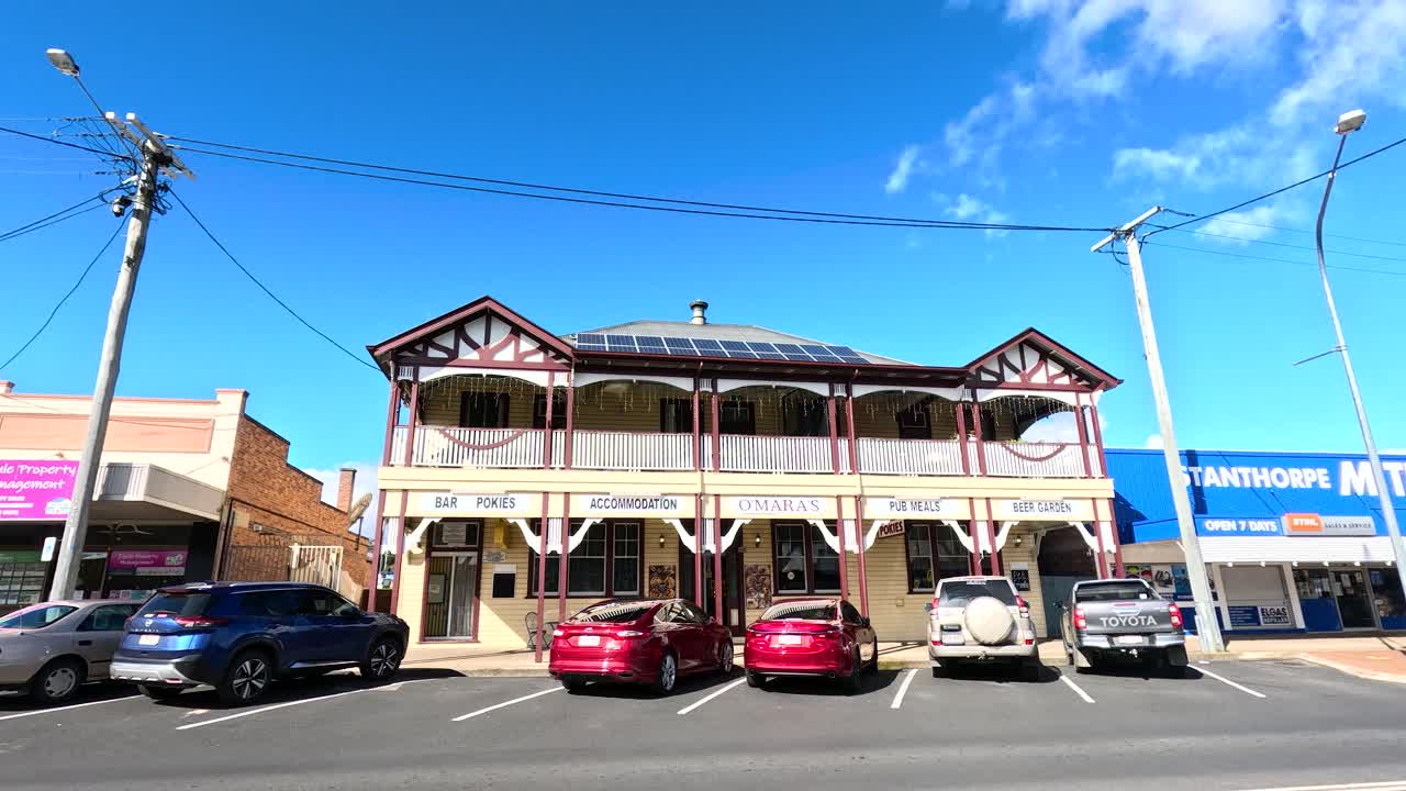 A white SUV passes in front of a traditional two-story Australian pub on a sunny day in Stanthorpe, Queensland. Static wide shot, bright daylight