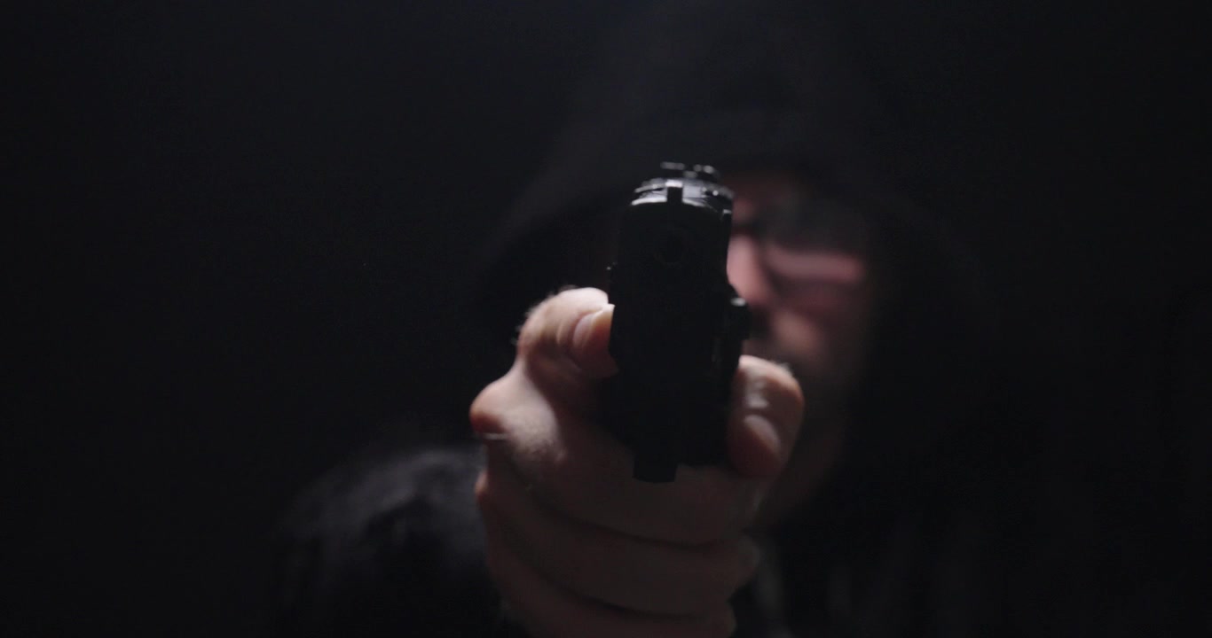 Homem encapuzado apontando uma pistola diretamente para a câmera em um ambiente escuro, representando crime, ameaça pessoal, perigo, assalto à mão armada e conceitos de segurança