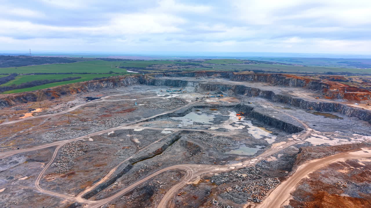 Quarry with machines and terrain shifts. Workers operate machinery on a large quarry site during cloudy weather. The ground shows signs of excavation and change