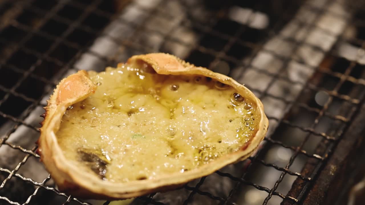 Close-up of a crab shell filled with bubbling sauce, grilling on a metal grate.