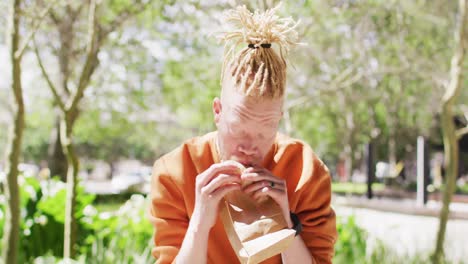Albino-african-american-man-with-dreadlocks-sitting-in-park-with-bike-eating-sandwich