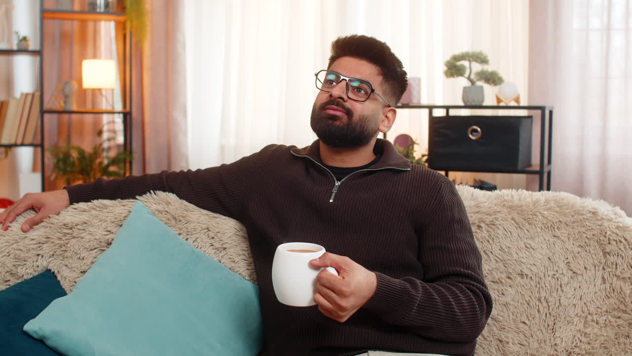 Indian man at home sofa enjoys tea or coffee after long day smiling and relaxing in peaceful mood