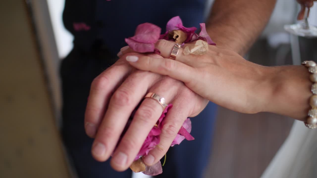 Slow motion close-up of bride and groom holding hands with weddings rings and falling flower petals