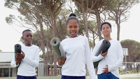 multiracial students holding yoga mats on field, smiling and ready for school activity