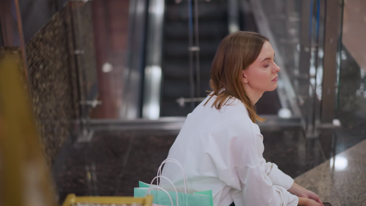 쇼핑몰의 벤치에 앉아있는 젊은 여성, 그녀의 에 다채로운 쇼핑 가방, 배경에 에스컬레이터, 사려 깊은 표정, 축제 분위기, 그리고 유리 레일링에 미묘한 빛 반사