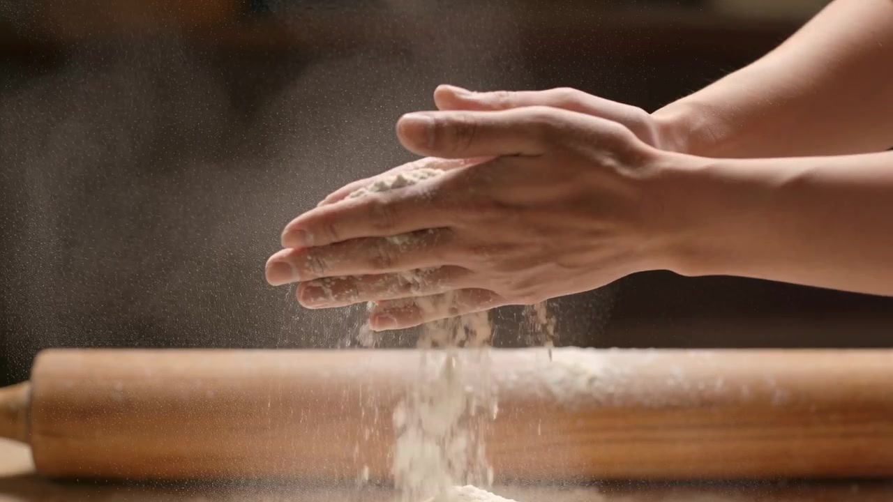 Mani di fornaio con maniche corte che versano farina su un bancone di legno per la preparazione dell'impasto, con un mattarello.