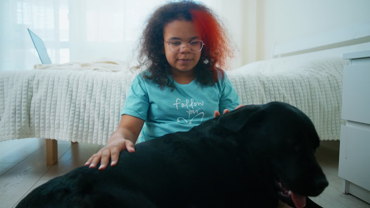 Young girl tenderly hugging black Labrador, sharing warm moment of affection while sitting on bedroom floor near bed, expressing deep bond between child and pet