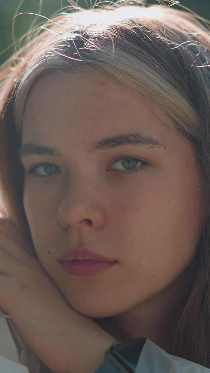 primer plano de una mujer joven descansando su barbilla en su mano, con una expresión reflexiva y la luz del sol suave destacando su cara, la vegetación borrosa en el fondo