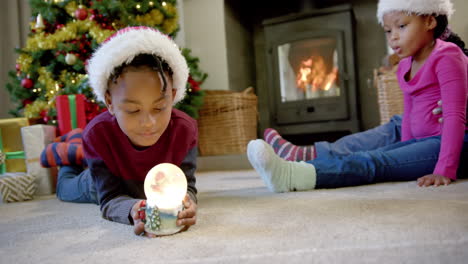 Hijo-Afroamericano-Con-Sombrero-De-Navidad-Sosteniendo-Un-Globo-De-Nieve-Brillante,-Familia-En-El-Fondo,-Cámara-Lenta