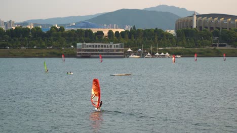 Menschen,-Die-Bei-Sonnenuntergang-Auf-Dem-Han-Fluss-In-Der-Nähe-Des-Ttukseom-Parks,-Des-Jamsil-Sportkomplexes-Oder-Der-Jamsil-Arena-Und-Entfernter-Berge-Im-Hintergrund-Windsurfen