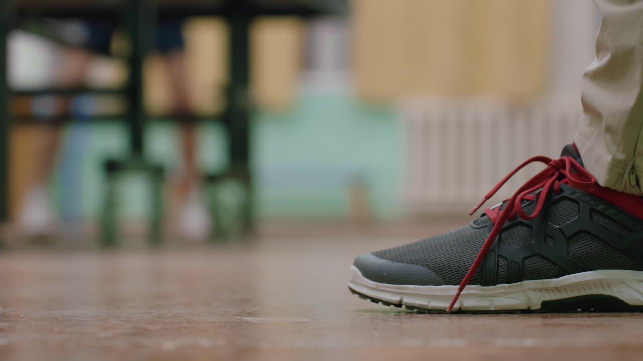 Close up of young tennis player wearing dark sneaker with red laces and beige pants standing on indoor court, detail shot highlighting footwear, stability, and preparation during sports activity