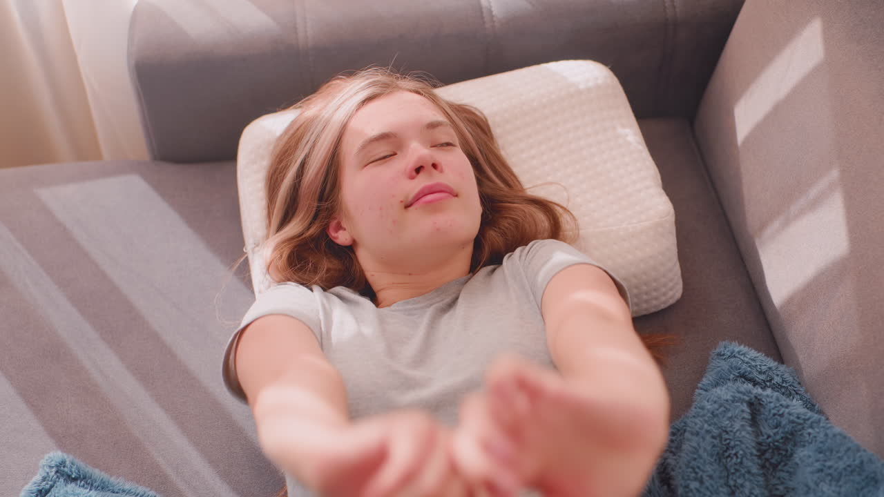 Close up of elegant lady waking up finally stretching herself on couch, resting with soft blanket and pillow as morning sunlight streams through curtains creating cozy peaceful atmosphere
