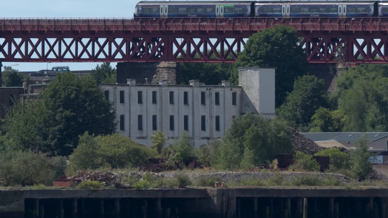 Modern passenger train travels across iconic red railway bridge above lush greenery, daylight, wide shot