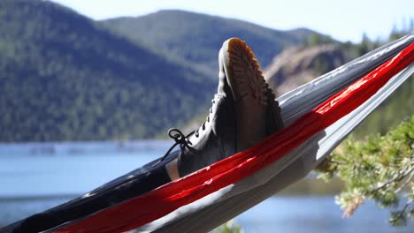 Close-up-of-boots,-crossed-legs-relaxing-in-swinging-hammock-by-lake