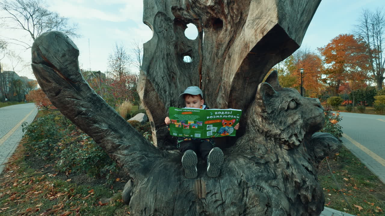 Male kid sitting in the wooden chair cut from tree. Lovely baby holding a book. Autumn park at backdrop.