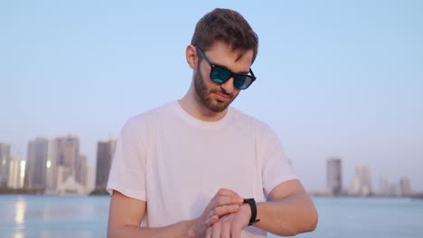 happy handsome man in sunglasses and white t-shirt uses a watch watch looks and presses his finger on the screen standing on the waterfront in summer against the city and buildings