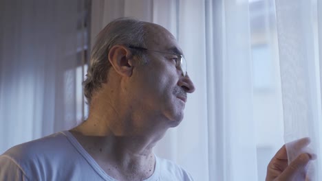 un viejo infeliz mirando por la ventana de la sala de estar en casa se quita las gafas.