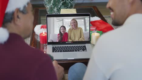 Biracial-father-and-son-with-santa-hats-using-laptop-for-christmas-video-call-with-family-on-screen