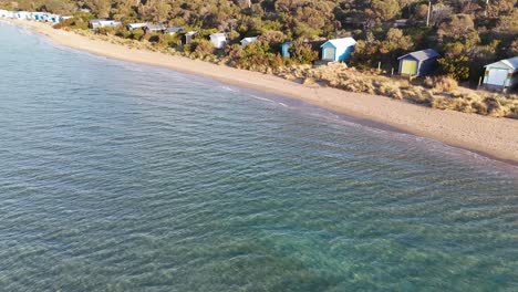 Drone glides above sandy beach, turquoise water, and vibrant bathing boxes in golden sunlight