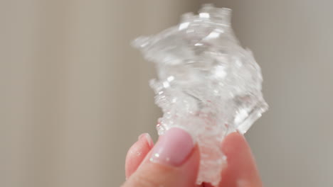 close up of hand with pink polished nails shaking transparent rubber-like soluble material, water droplets visible on fingers, natural daylight highlighting texture against blurred indoor background