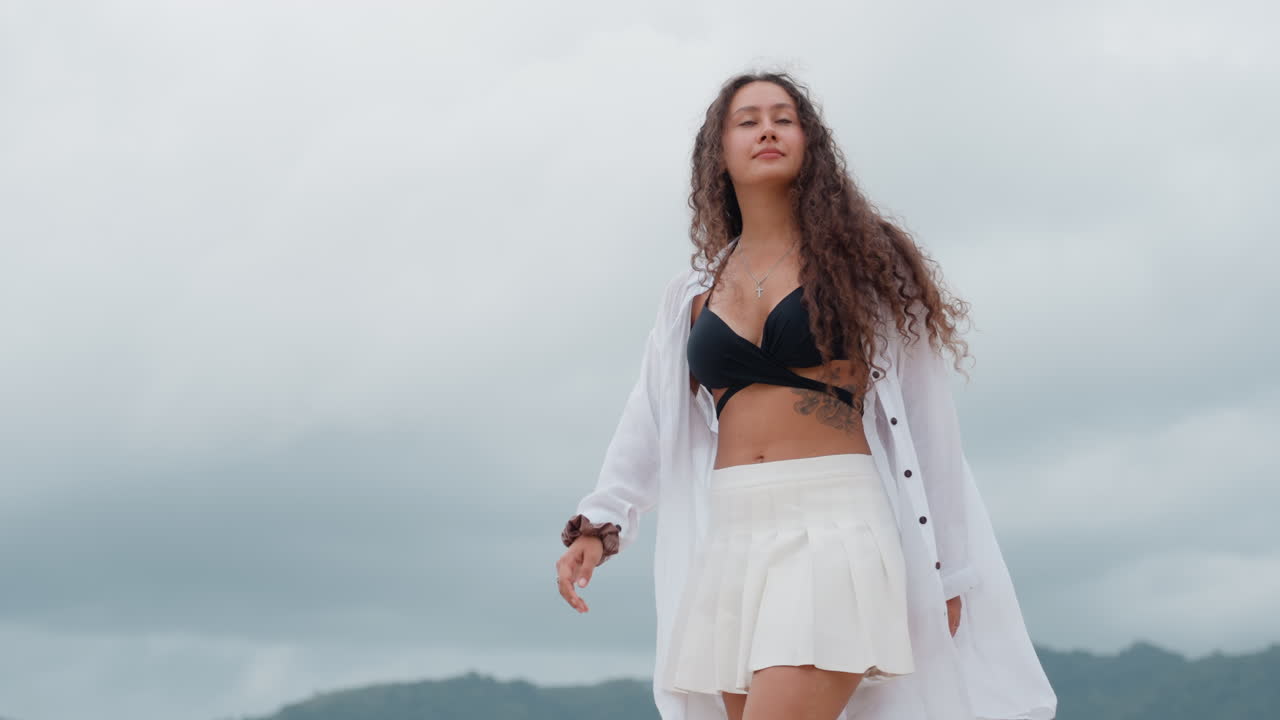 Posa di donna in spiaggia. Signora in riva al mare con capelli fluenti. Donna in piedi accanto all'oceano calmo con colline nebbiose dietro. Serena silhouette femminile vicino alla riva tra tempo coperto e brezza leggera.
