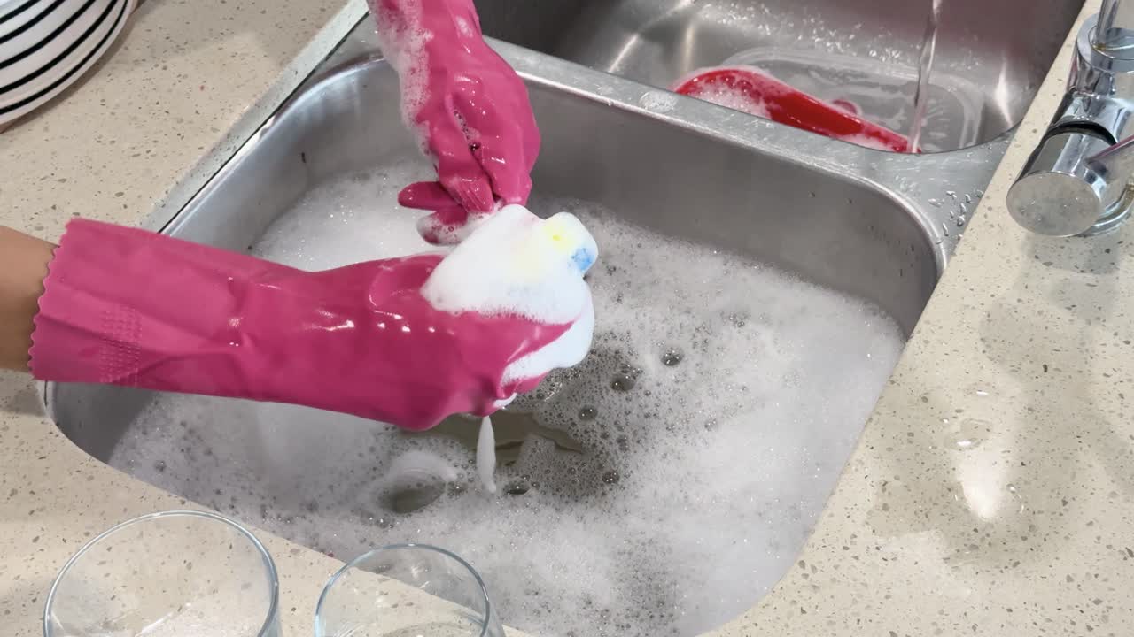 Person wearing pink gloves scrubs a metal spoon with a sponge in a modern kitchen sink filled with soapy water, under bright lighting