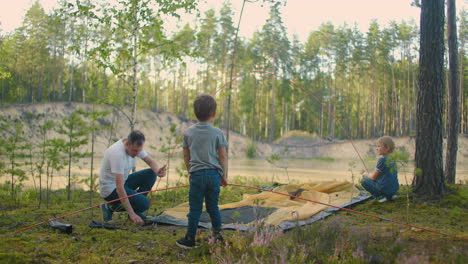 the young man and two young boys together set up a tent camp for the night during the campaign. fatherhood and a happy childhood. father and sons put up a tent together in the woods