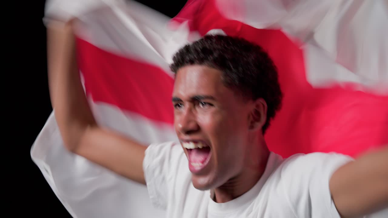 Uomo afroamericano in maglietta bianca che alza e sventola una bandiera con una croce rossa in studio, celebrando.