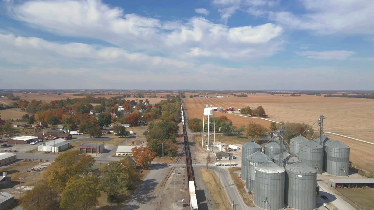 Drohnenaufnahme, die einem Zug mit Waggons folgt, der auf Eisenbahnschienen durch eine ländliche Gegend mit Silos und einem Wasserturm fährt