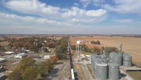 drone shot following a train with carriages travels on railroad tracks amonga farm zone with silos and a water tower