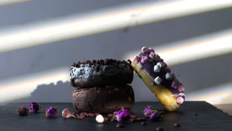 a hand with pink nails lifts a donut topped with chocolate cookie crumbles from a stacked trio of colorful donuts. the slate surface, soft shadows, and scattered rosebuds create a cozy, moody dessert.