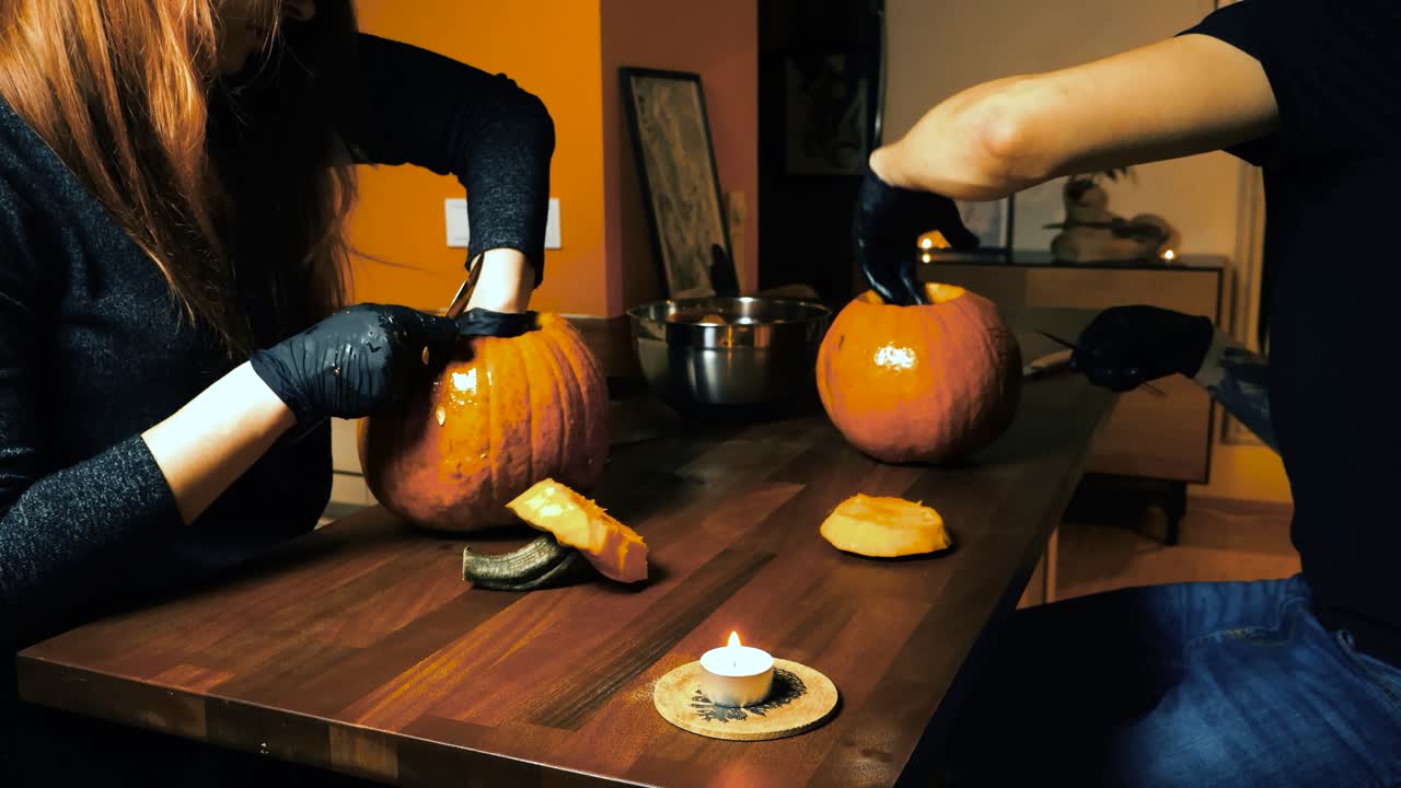 Two people a man and a woman carving out Halloween pumpkins with sharp knifes while sitting at a table where candles are burning. Video is done in time lapse by revealing the finished pumpkins at end.