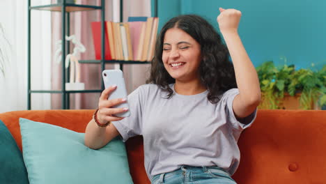 Indian-Arabian-woman-at-home-use-smartphone-celebrating-success-victory-winning-play-online-game