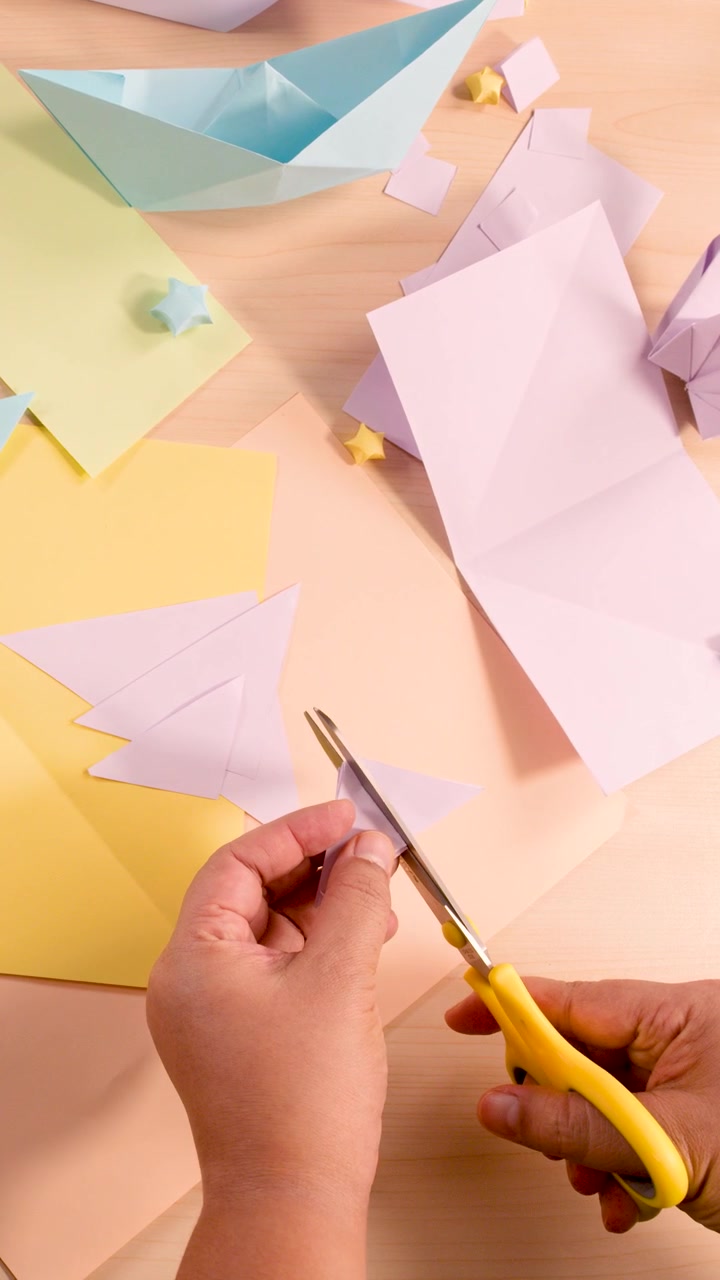 Mani adulte piegano e tagliano carta pastello per origami su un tavolo ben illuminato, vista dall'alto.