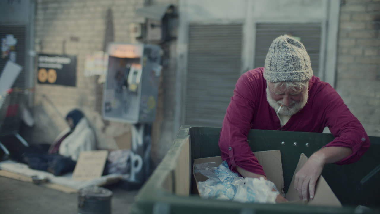 Senior Homeless Man Smoking and Rummaging through Trash Bin