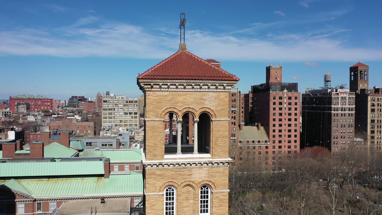 An aerial view over the bell tower of a church in Greenwich Village, NYC. The drone camera orbit counterclockwise around the red roof tower on a sunny day. There are other buildings in the background.